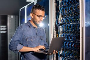 A young man holding an open gray laptop stands in front of a data center.