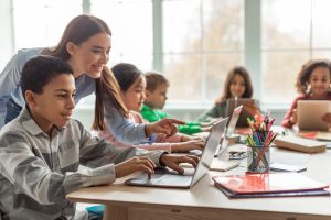 A teacher leans over her students and points to the electronic learning device which is being used for teaching.